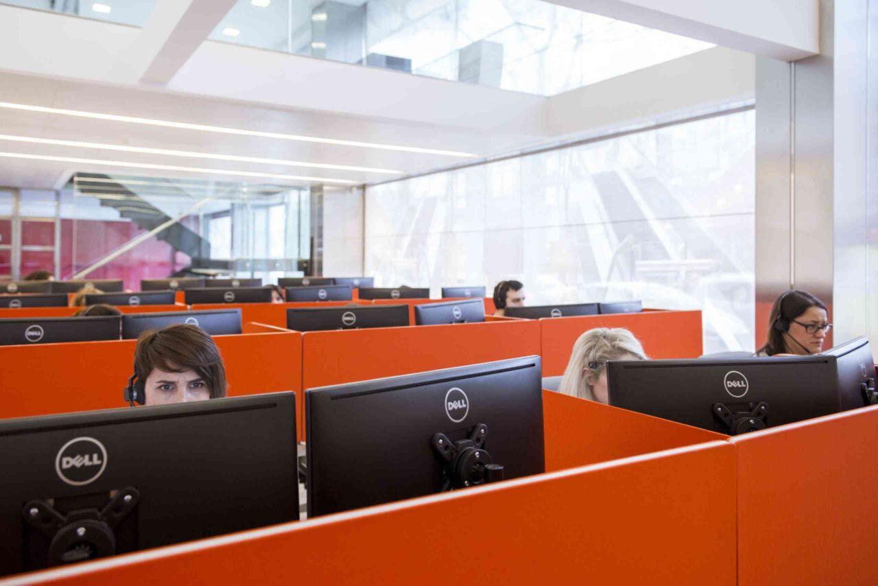 Centre d'atenció al client amb diversos empleats de FAIN treballant en cubicles taronges davant de monitors Dell, usant auriculars amb micròfon en una oficina moderna i lluminosa.